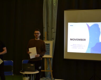 Zbliżenie. Dwóch mężczyzn stoi przy ekranie. Na ekranie napis Movember. Mężczyźni w dłoniach trzymają kartki.
