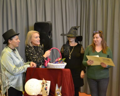 Zbliżenie. Cztery kobiety stoją przy stoliku mikrofonem. Jedna kobieta trzyma koszyk, druga papierową teczkę.