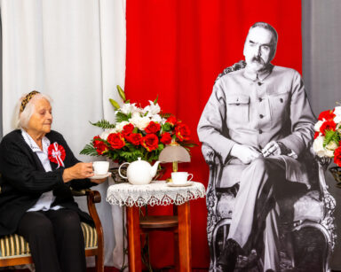 Zbliżenie. Kobieta siedzi przy stoliku z Marszałkiem Piłsudskim. Obok biło czerwone kwiaty. W tle flaga Polski.
