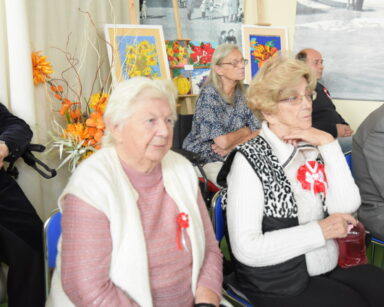 Sala. Seniorzy siedzą na krzesłach. Kobiety mają przypięty kotylion. W tle obrazy w kwity.
