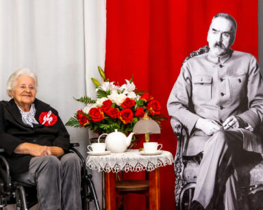 Zbliżenie. Kobieta siedzi przy stoliku z Marszałkiem Piłsudskim. Obok biło czerwone kwiaty. W tle flaga Polski.