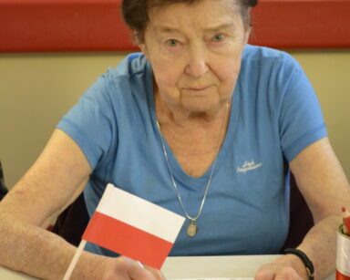 Zbliżenie. Kobieta koloruje Herb Polski. Na stole w dzbanuszku czerwona róża i mała flaga Polski.
