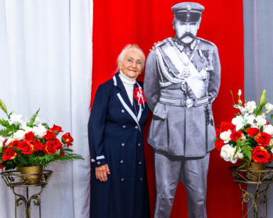 Zbliżenie. Kobieta pozuje do zdjęcia z postacią Piłsudskiego. Obok biało czerwone kwiaty. W tle flaga Polski.
