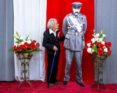 Zbliżenie. Kobieta pozuje do zdjęcia z Piłsudskim. Kobieta przygląda się Marszałkowi. Obok biało czerwone kwiaty.