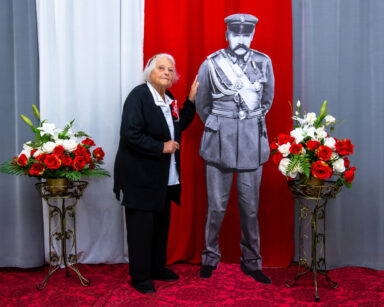 Zbliżenie. Kobieta pozuje do zdjęcia. Obok postać Marszałka Piłsudskiego. W tle flaga Polski.