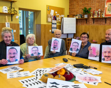 Sala. Grupa ludzi trzyma w dłoniach zdjęcia mężczyzn z domalowanymi wąsami. Na stole kartki z różnymi kształtami wąsów.