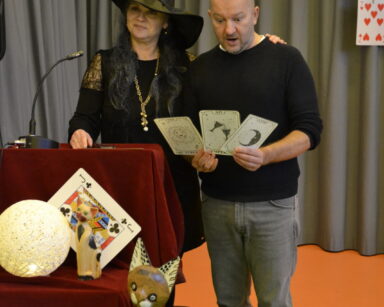 Zbliżenie. Kobieta w kapeluszu stoi koło mężczyzny. Mężczyzna trzyma w dłoni karty. Obok stoją kule i drewniany kot.