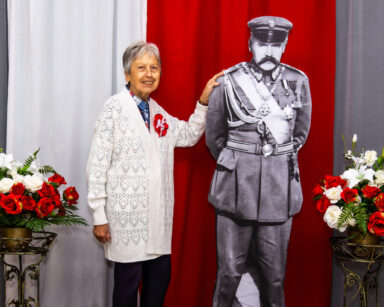 Zbliżenie. Uśmiechnięta kobieta pozuje do zdjęcia z Piłsudskim. Obok bukiety kwiatów. W tle flaga Polski.