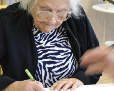 Zbliżenie. Seniorka siedzi przy stole, trzyma w ręku zieloną kredkę. Koloruje obrazek. Obok leżą kolorowe kredki.