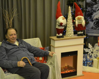Zbliżenie. Senior siedzi na kanapie. W ręku trzyma laskę. Obok kominek na nim ozdoby świąteczne. Obok stoją prezenty.