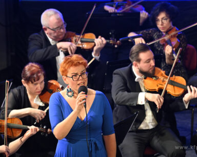 Zbliżenie. Kobieta w niebieskiej sukience trzyma mikrofon. W tle grupa muzyków ze skrzypcami w dłoniach.