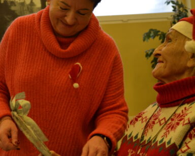 Zbliżenie. Dwie kobiety patrzą na ozdobę świąteczną. Kobiety ubrane są w czerwone swetry. Obie panie uśmiechają się.