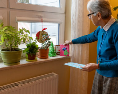 Zbliżenie. Seniorka w niebieskim swetrze układa na parapecie kartki świąteczne. Obok na oknie stoją rośliny.