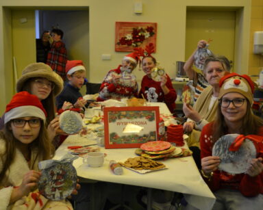 Sala. Grupa seniorów i dzieci siedzi przy stole. W rękach trzymają własnoręcznie zrobione ozdoby świąteczne.