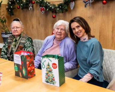 Zbliżenie. Młoda kobieta i dwie seniorki pozują do zdjęcia Przed nimi stół. Na stole stoją świąteczne torebki z prezentami.