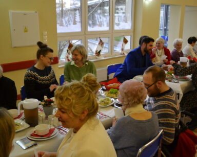 Sala. Grupa ludzi siedzi przy stole. Na stole wigilijne potrawy. W tle ozdoby świąteczne na oknach.