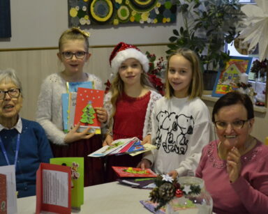 Sala. Dwie seniorki i trzy dziewczynki pozują do zdjęcia. Wszyscy w dłoniach mają świąteczne kartki.