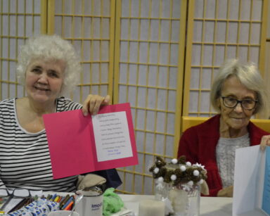 Sala. Dwie seniorki podnoszą do zdjęcia świąteczne kartki. Na stole przed kobietami ozdoby i kredki.