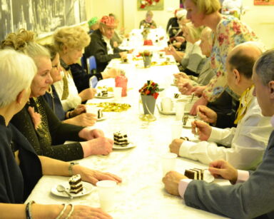 Sala. Grupa osób siedzi przy stole. Na stole stoją talerzyki z ciastem i kubeczki. W tle na ścianach zdjęcia.