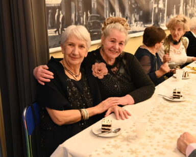 Zbliżenie. Dwie kobiety przy stole pozują do zdjęcia. Ubrane są w czarne suknie. W tle seniorzy siedzą przy stole.