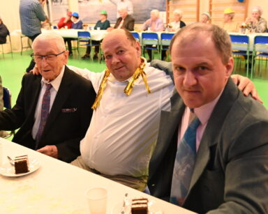 Zbliżenie. Trzech mężczyzn siedzi przy stole i pozuje do zdjęcia. Na stole stoją talerzyki z ciastem.