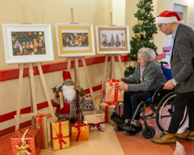 Korytarz. Seniorka na wózku trzyma w ręku prezent. Obok stoi mężczyzna. W tle zdjęcia , prezenty i choinka.