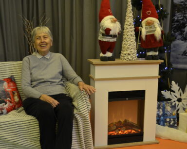 Zbliżenie. Kobieta siedzi na kanapie. Obok kominek, prezenty, Mikołaje i mała biała choinka.