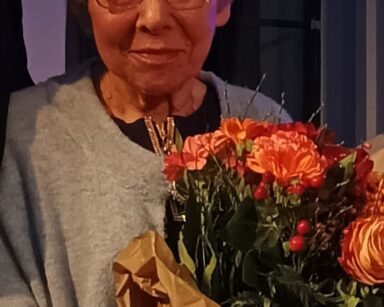 Zbliżenie. Starsza kobieta w okularach z bukietem kwiatów w dłoni pozuje do zdjęcia. Kobieta uśmiecha się.