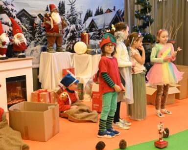 Scena. Grupa dzieci robi przedstawienie. W tle kominek, prezenty, figurki Mikołaja, choinki i zimowa sceneria.