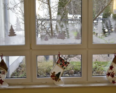 Zbliżenie. Okno. Na szybach namalowane choinki i śnieg. Do klamek przymocowane są świąteczne skarpety.