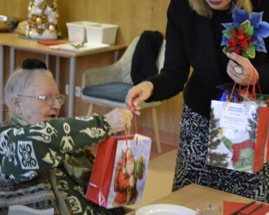 Sala. Kobieta z kwiatem w marynarce podaje prezent seniorce.