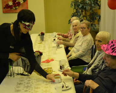 Sala. Kobieta w czarnej sukni i masce na twarzy stawia filiżankę z kawą na stół. Przy stole siedzą seniorzy.