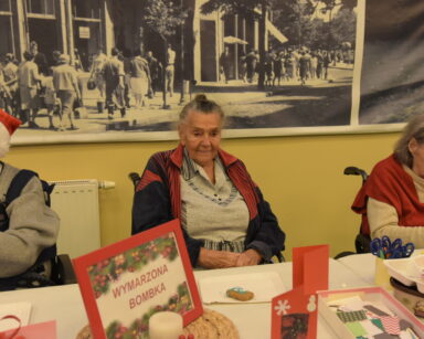 Zbliżenie. Trzej seniorzy siedzą przy stole. Na stole kartka z napisem Wymarzona bombka oraz materiały do dekoracji.