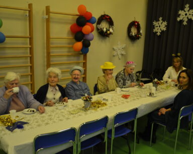Sala. Osiem osób siedzi przy stole. Na stole sylwestrowa maska ciasto, łyżeczki. Na ścianach balony, stroiki, śnieżynki.