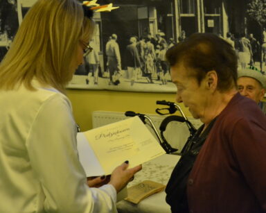 Zbliżenie. Kobieta przekazuje seniorce podziękowania w formie dyplomu. W tle na ścianie czarno białe zdjęcie.