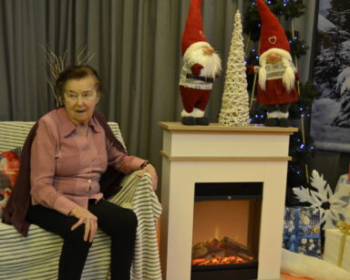 Zbliżenie. Kobieta w różowej bluzce siedzi na kanapie. Obok kominek, prezenty, dwa Mikołaje i biała choinka.