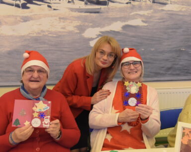Zbliżenie. Trzy kobiety pozują do zdjęcia. Dwie seniorki trzymają w dłoniach świąteczne kartki.