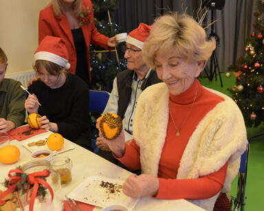 Zbliżenie. Uśmiechnięta seniorka trzyma w dłoni pomarańczę z goździkami. Obok przy stole siedzą dzieci.