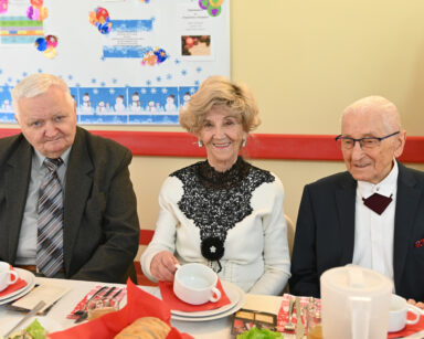 Zdjęcie. Trzech uśmiechniętych seniorów siedzi przy stole. W tle na tablicy świąteczne życzenia.
