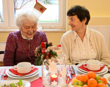 Zdjęcie. Dwie uśmiechnięte seniorki siedzą przy stole. Na stole zastawa świąteczna.