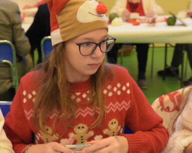 Zbliżenie. Młoda dziewczynka siedzi przy stole. Ubrana jest w czapkę Mikołaja i świąteczny sweter.