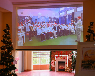 Zbliżenie. Ekran. Na ekranie grupa ludzi w czapkach Mikołaja. Przy ekranie choinki świąteczne, kominek i ozdoby.