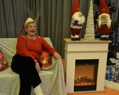 Zbliżenie. Uśmiechnięta kobieta w stroju Mikołaja siedzi na kanapie. Obok kominek, prezenty, choinka i dwa Mikołaje.