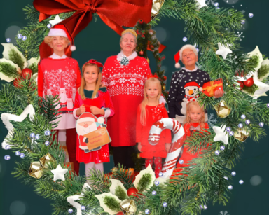 Zdjęcie. Świąteczny wieniec z czerwoną kokardą. W środku stoją seniorzy i małe dziewczynki. Napis Wesołych Świąt.