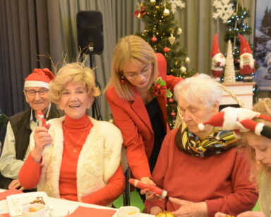 Zbliżenie. Uśmiechnięci seniorzy siedzą przy stole w ręku trzymają Mikołaja. Obok stoi kobieta i podaje czekoladkę.