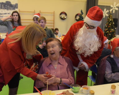 Zbliżenie. Uśmiechnięta seniorka odbiera czekoladę od kobiety z koszykiem. Obok stoi Mikołaj. W tle grupa ludzi.
