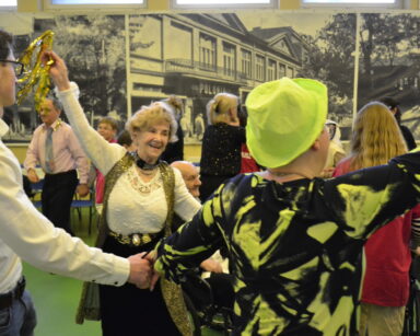 Sala. Grupa uśmiechniętych ludzi tańczy. Seniorka w ręku trzyma złote wstążki.