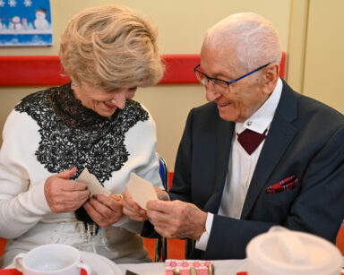 Zbliżenie. Para elegancko ubranych seniorów dzieli się opłatkiem. Seniorzy uśmiechają się, siedzą przy stole.
