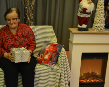 Zbliżenie. Seniorka trzyma prezent. Siedzi na kanapie. Obok kominek z ozdobami świątecznymi.