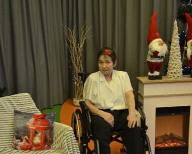 Zbliżenie. Kobieta siedzi na wózku inwalidzkim, na głowie ma czerwoną spinkę. Obok kominek, choinka i dwa Mikołaje.